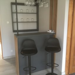 DARCO-USŁUGI MONTAŻOWE ZABIELSKI DARIUSZ - Nowoczesny barek z szarą ladą, dwoma czarnymi hokerami i metalową półką na kieliszki. Na półce dekoracyjne szklanki. Całość w jasnym wnętrzu z drewnianymi akcentami.