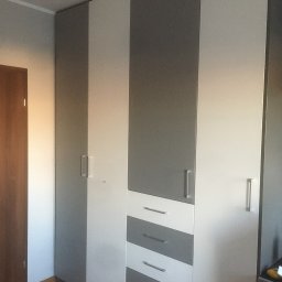 DARCO-USŁUGI MONTAŻOWE ZABIELSKI DARIUSZ - Wysoka szafa z szarymi i białymi frontami, z widocznymi metalowymi uchwytami i szufladami, stoi w pokoju z drewnianą podłogą. Minimalistyczny design.