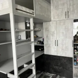 Funkcjonalna zabudowa pokoju młodzieżowego z łóżkiem na antresoli, biurkiem i szafami. Biało-szara kolorystyka, nowoczesny design. Widoczna drabinka i półki.