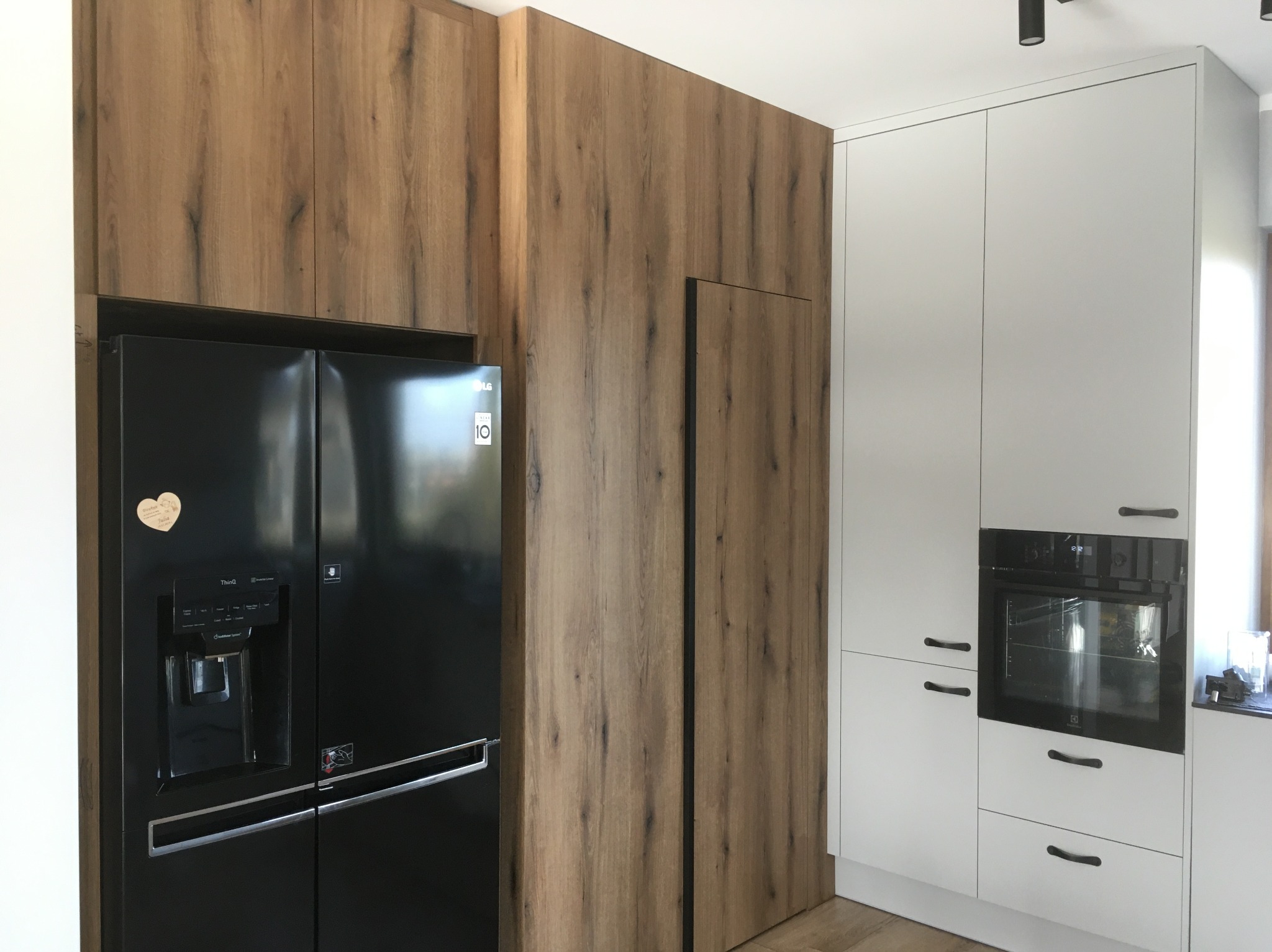 Nowoczesna kuchnia z zabudową drewnopodobną, integrującą czarną lodówkę side-by-side. Białe szafki z piekarnikiem i minimalistycznymi uchwytami dopełniają aranżację.