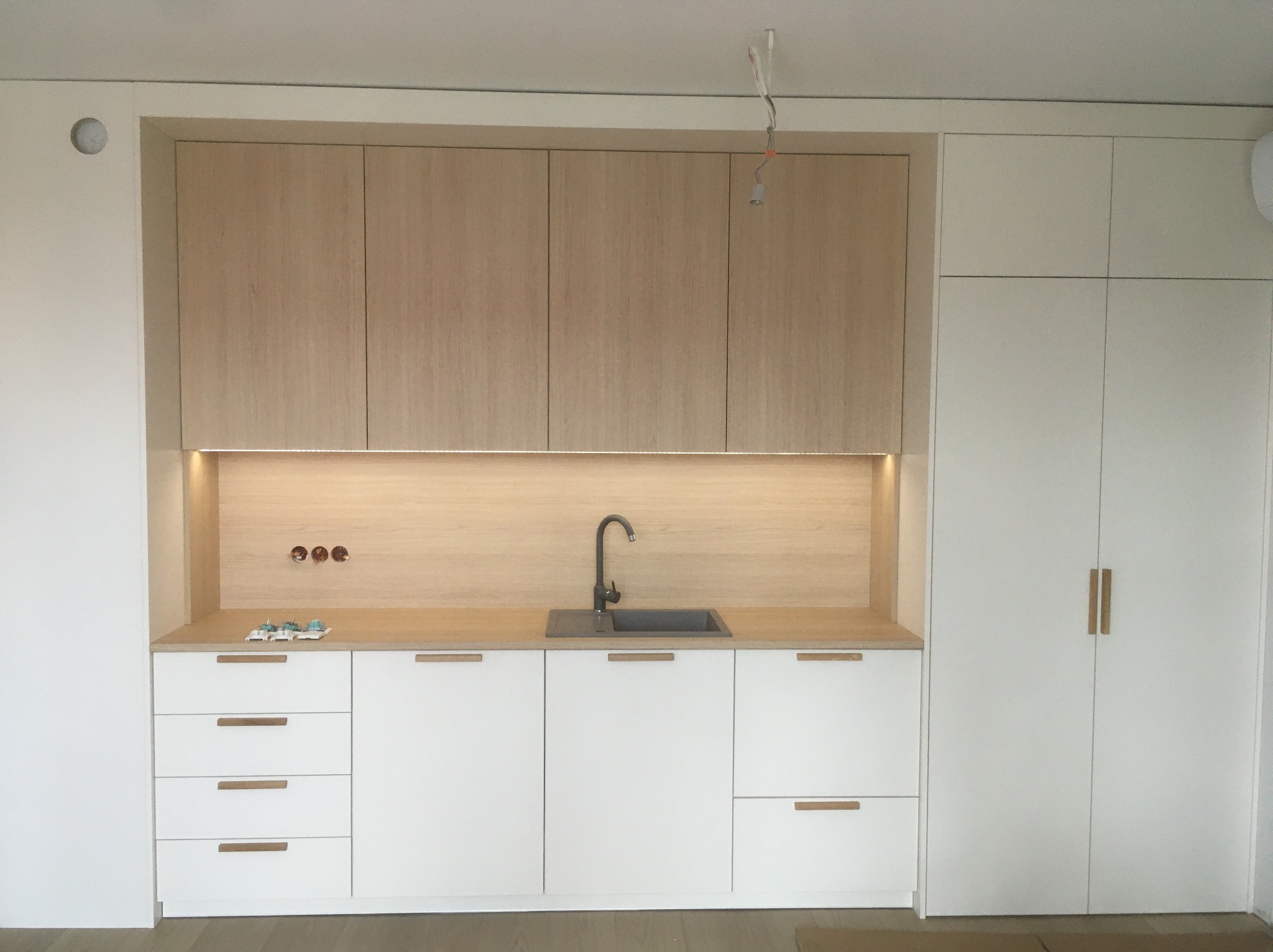 Jasna kuchnia na wymiar z drewnianymi akcentami: białe fronty, blat i szafki górne w odcieniu drewna, minimalistyczny zlew i bateria. Widoczne gniazdka i niedokończone oświetlenie.