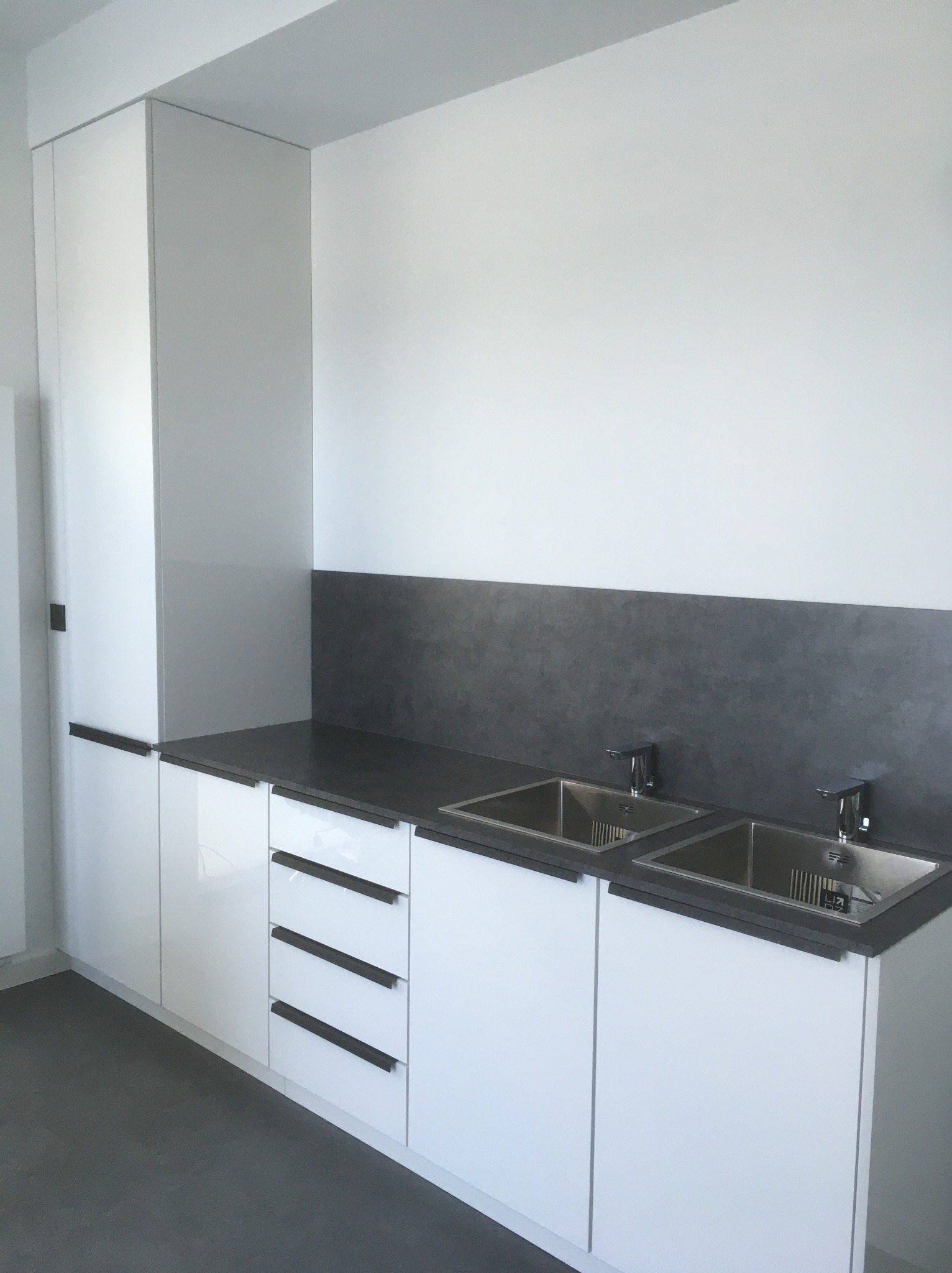 Minimalistyczna, biała zabudowa kuchenna z ciemnym blatem i dwoma zlewozmywakami. Nowoczesne, długie uchwyty podkreślają geometrię szafek.