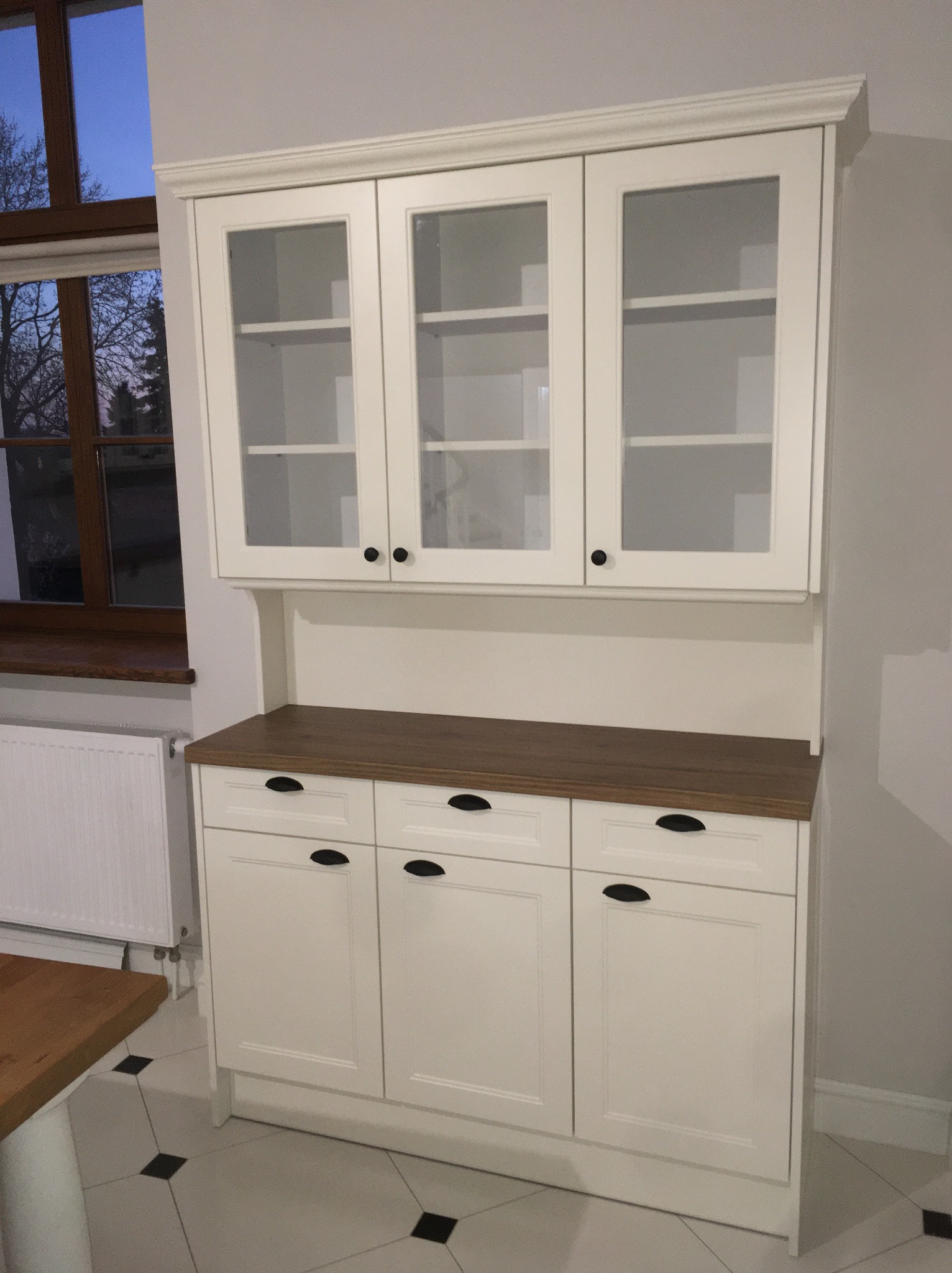 Biały kredens kuchenny z przeszkloną nadstawką i drewnianym blatem, widok z boku, czarne uchwyty, okno w tle.