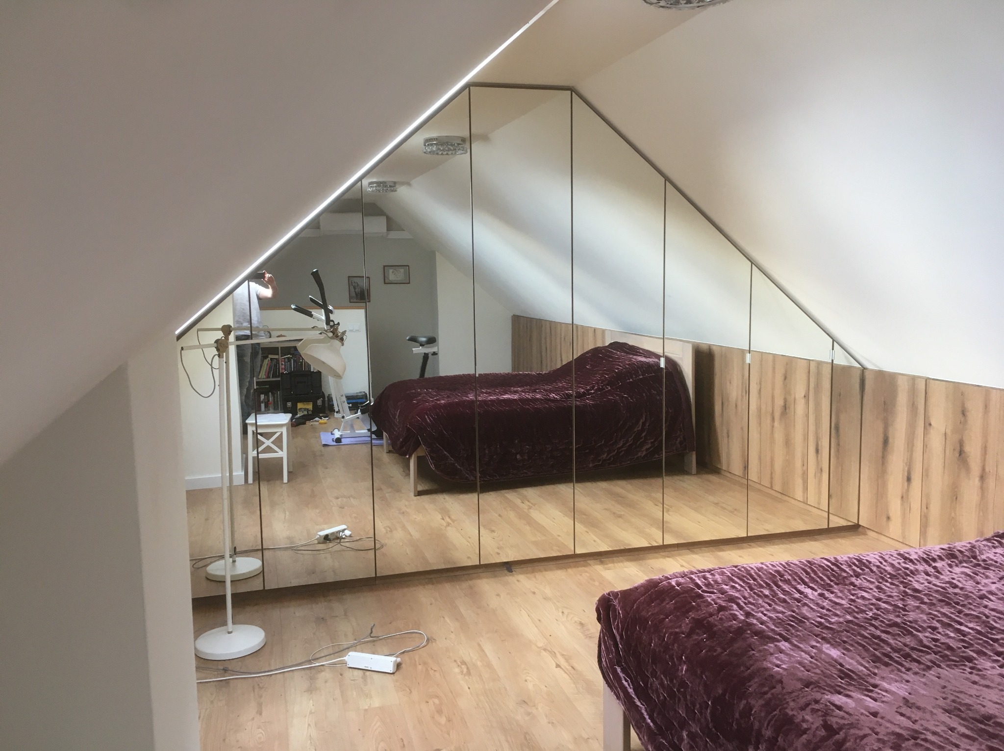 Szafa wnękowa z lustrzanymi drzwiami w pokoju na poddaszu, odbicie wnętrza pokoju. Widoczna część łóżka z bordową narzutą i drewniane wykończenie szafy.