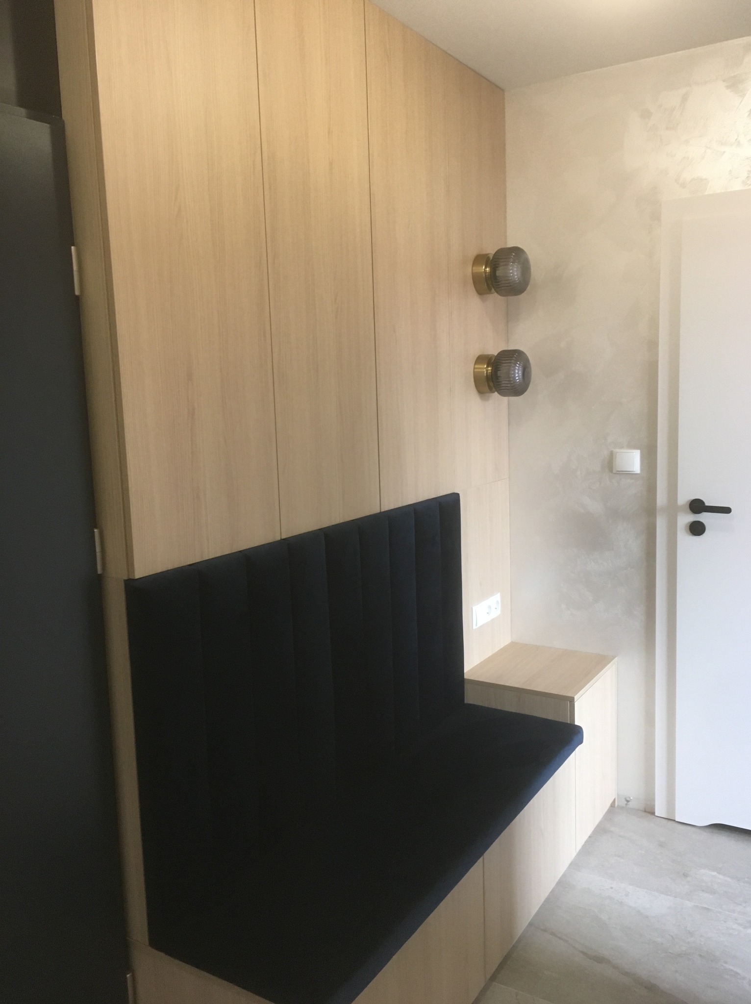 Jasna szafa wnękowa z siedziskiem obitym granatowym materiałem. Dwa szklane kinkiety na ścianie. Minimalistyczny design, jasne drewno, gładkie fronty.