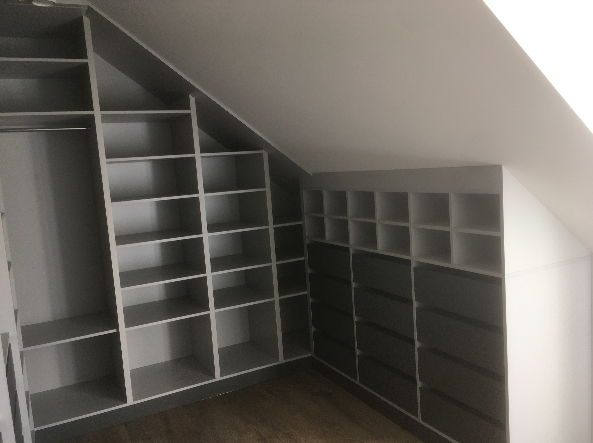 Szara szafa wnękowa z licznymi półkami i szufladami, dopasowana do skosu dachu. Widoczne drewniane panele podłogowe.