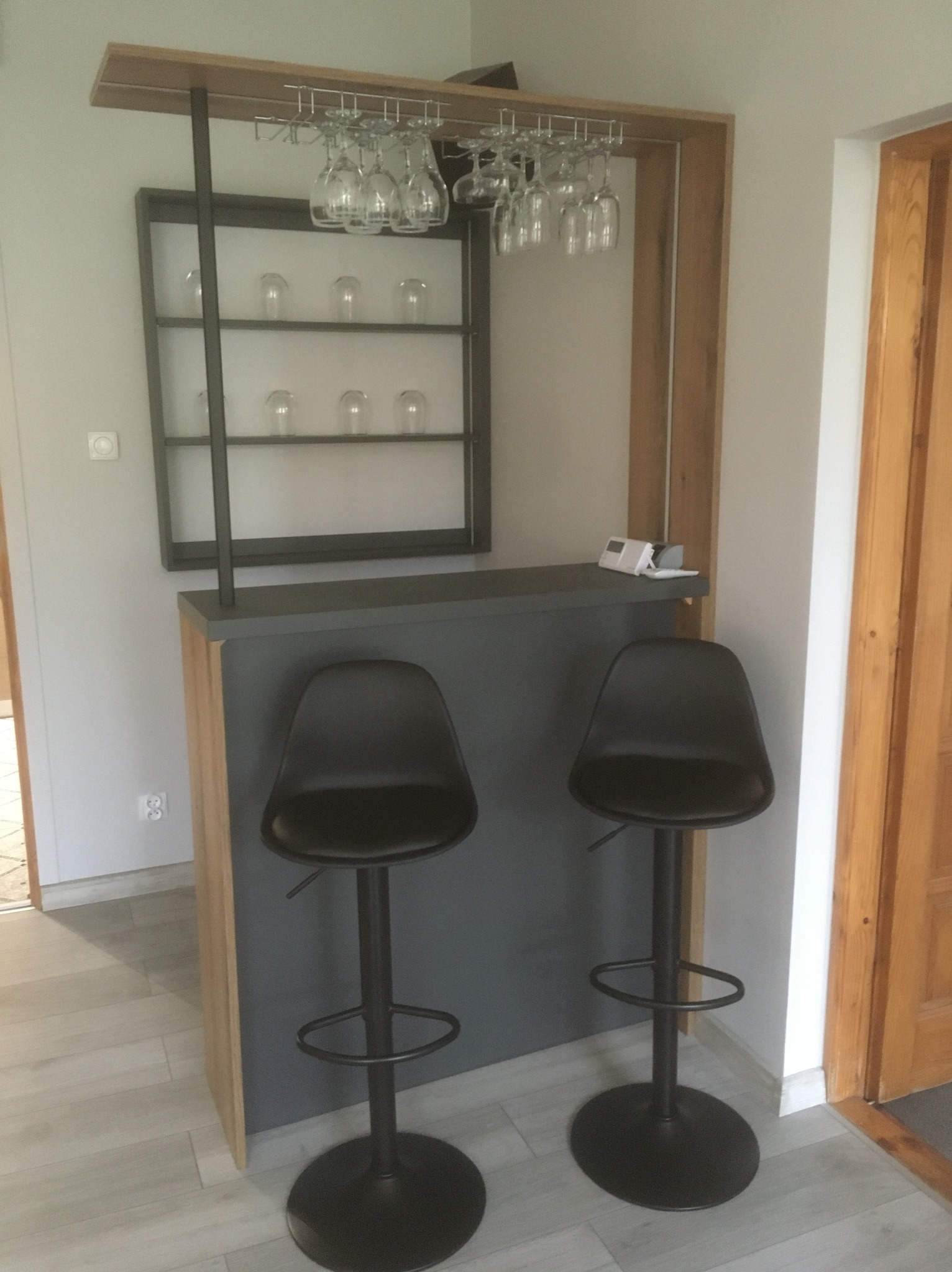 Nowoczesny barek z szarą ladą, dwoma czarnymi hokerami i metalową półką na kieliszki. Na półce dekoracyjne szklanki. Całość w jasnym wnętrzu z drewnianymi akcentami.