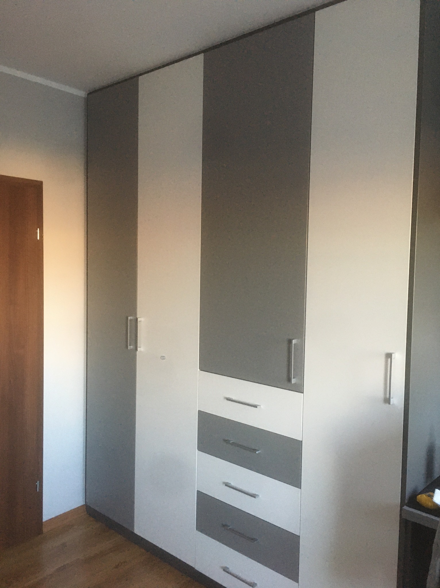 Wysoka szafa z szarymi i białymi frontami, z widocznymi metalowymi uchwytami i szufladami, stoi w pokoju z drewnianą podłogą. Minimalistyczny design.