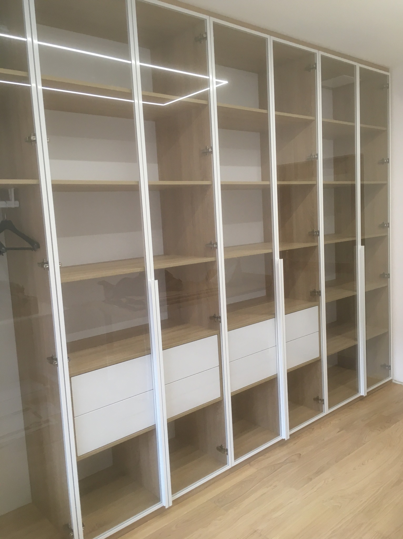 Wysoka szafa wnękowa z przeszklonymi drzwiami, oświetleniem LED i białymi szufladami, ukazana pod kątem, podkreślającym jej głębokość i nowoczesny design.