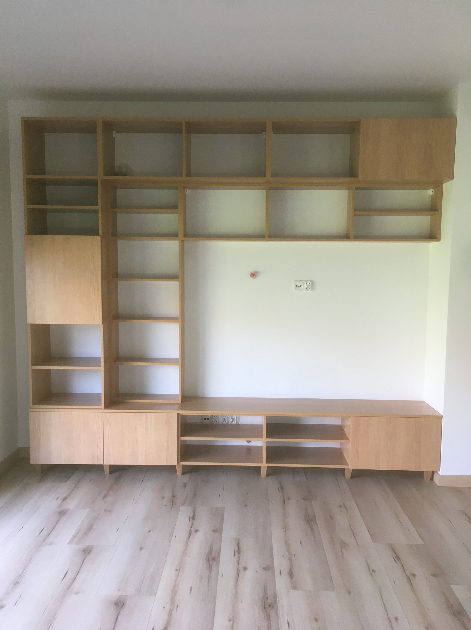 Jasna, drewniana zabudowa meblowa na ścianie z otwartymi półkami i szafkami, komponująca się z podłogą w odcieniach drewna. Minimalistyczny design, widoczne gniazdka elektryczne.