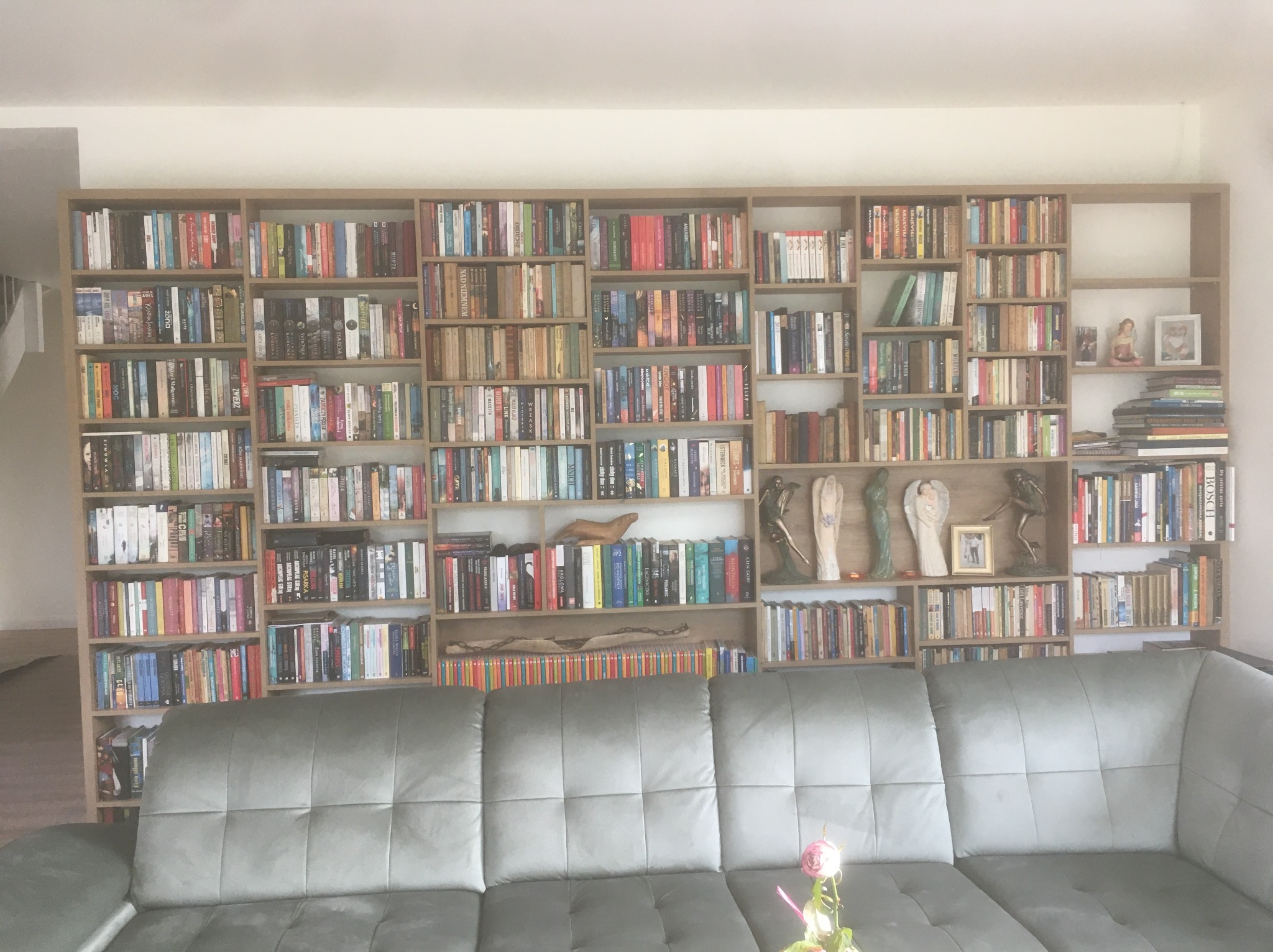 Duża, drewniana biblioteczka na całą ścianę, wypełniona książkami. Na półkach dekoracje i ramki ze zdjęciami. Na pierwszym planie szara kanapa z kwiatkiem.