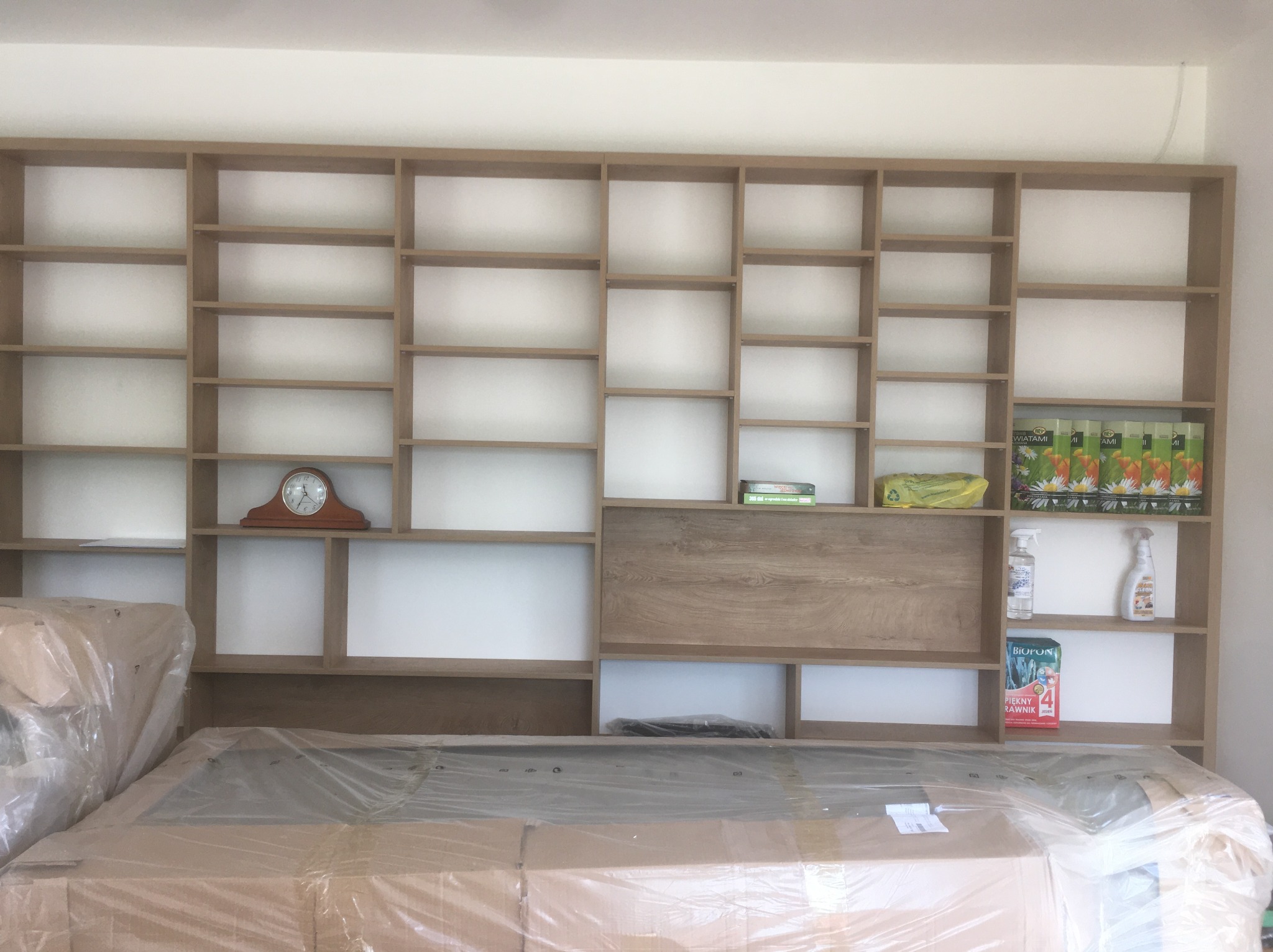 Jasna, drewniana szafa wnękowa z półkami o różnej wielkości, częściowo wypełniona przedmiotami, z blatem zabezpieczonym folią ochronną.