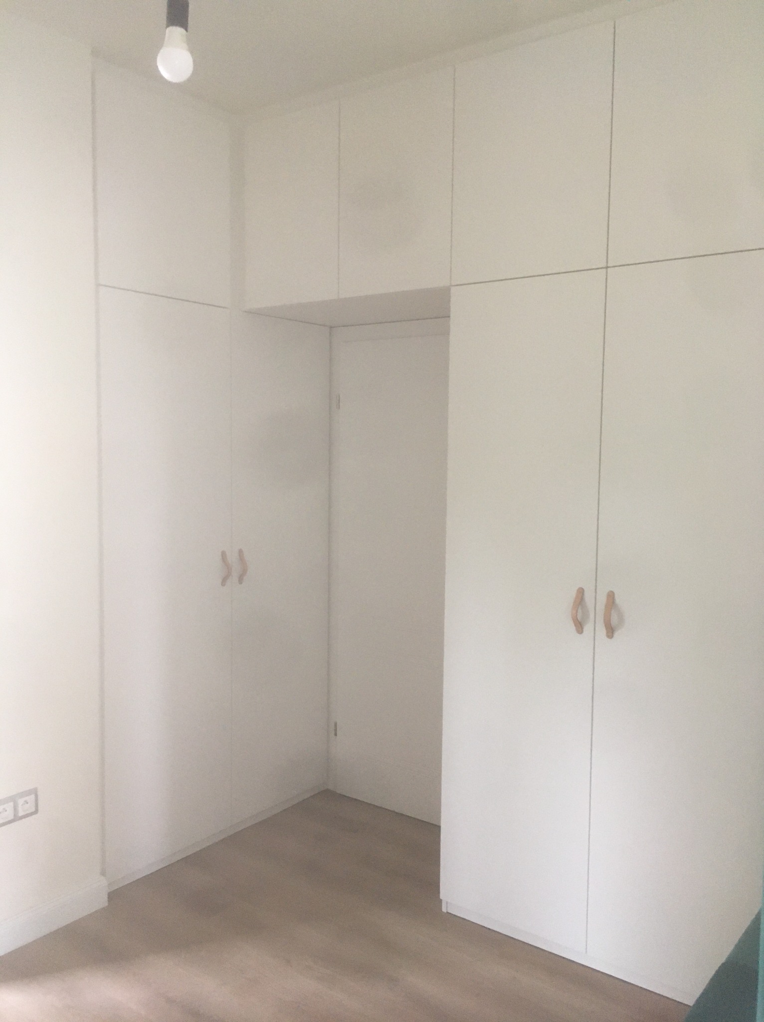 Biała zabudowa wnękowa z drewnianymi uchwytami, dopasowana do wysokości pomieszczenia, z widoczną przestrzenią na drzwi. Minimalistyczny design.