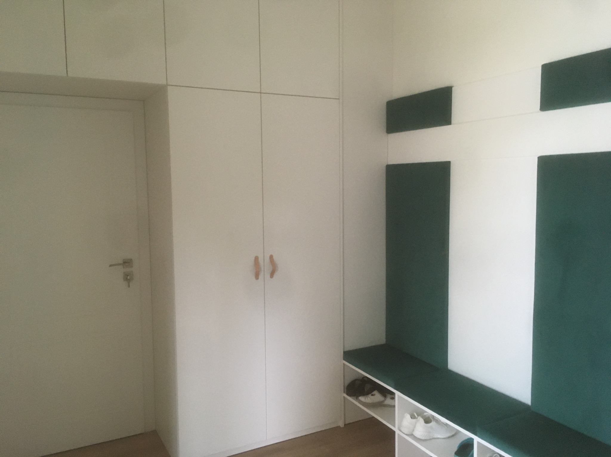 Biała szafa wnękowa z siedziskiem i półkami na buty, minimalistyczny design, drewniane uchwyty, zielone panele dekoracyjne na ścianie.