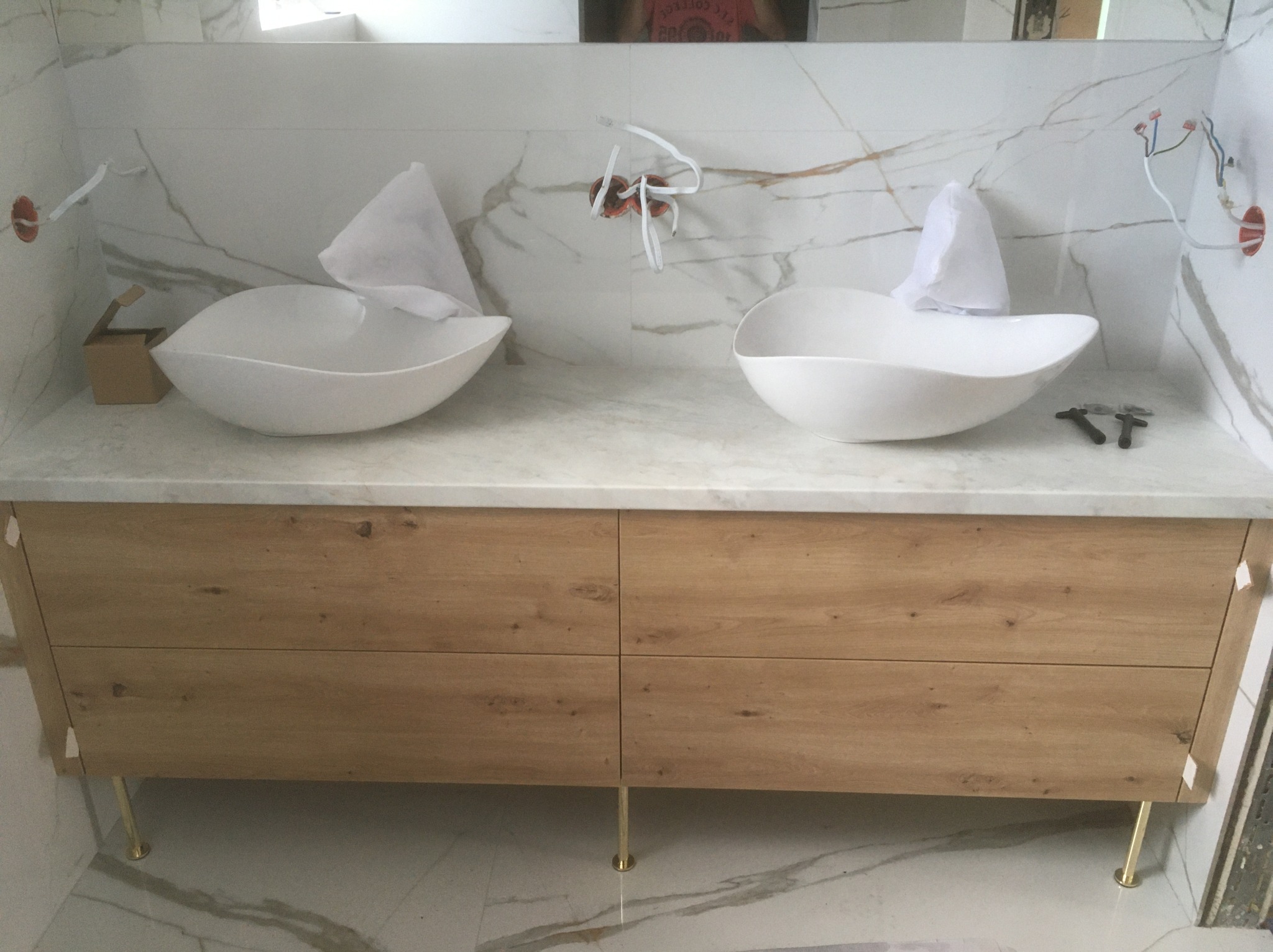 Łazienka z dwiema owalnymi umywalkami nablatowymi na drewnianej szafce z marmurowym blatem i złotymi nóżkami, w trakcie montażu instalacji elektrycznej.