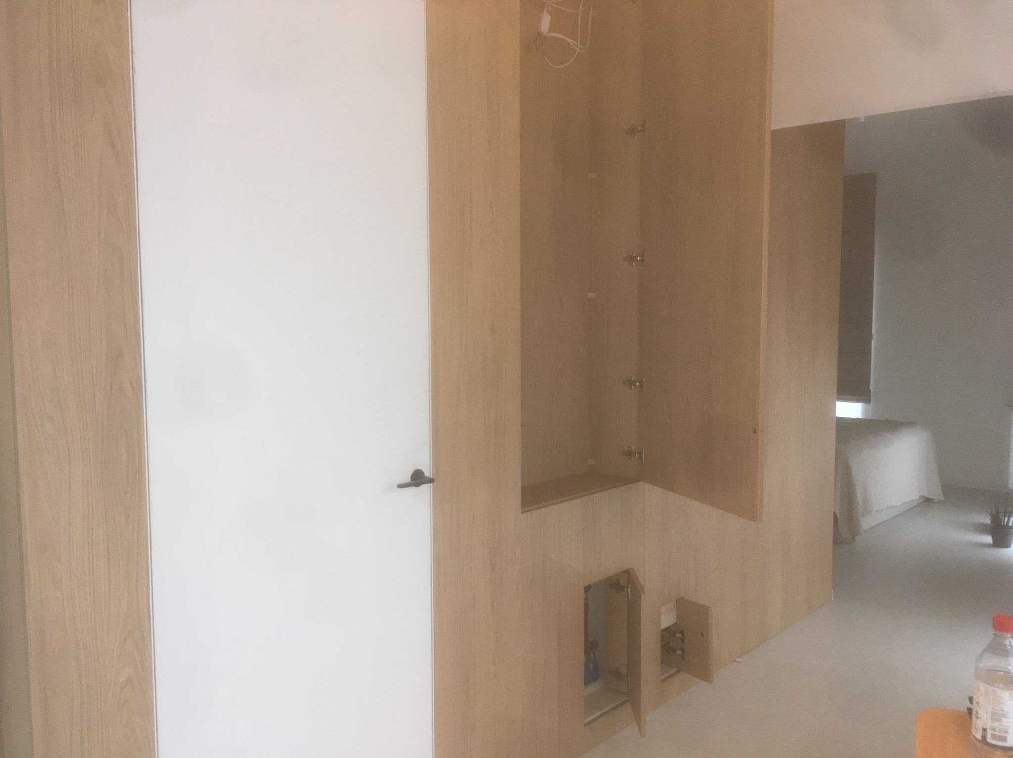 Wnętrze szafy wnękowej z jasnego drewna, z otwartymi drzwiczkami w dolnej części. Obok białe drzwi z czarną klamką. W tle widoczny fragment sypialni.