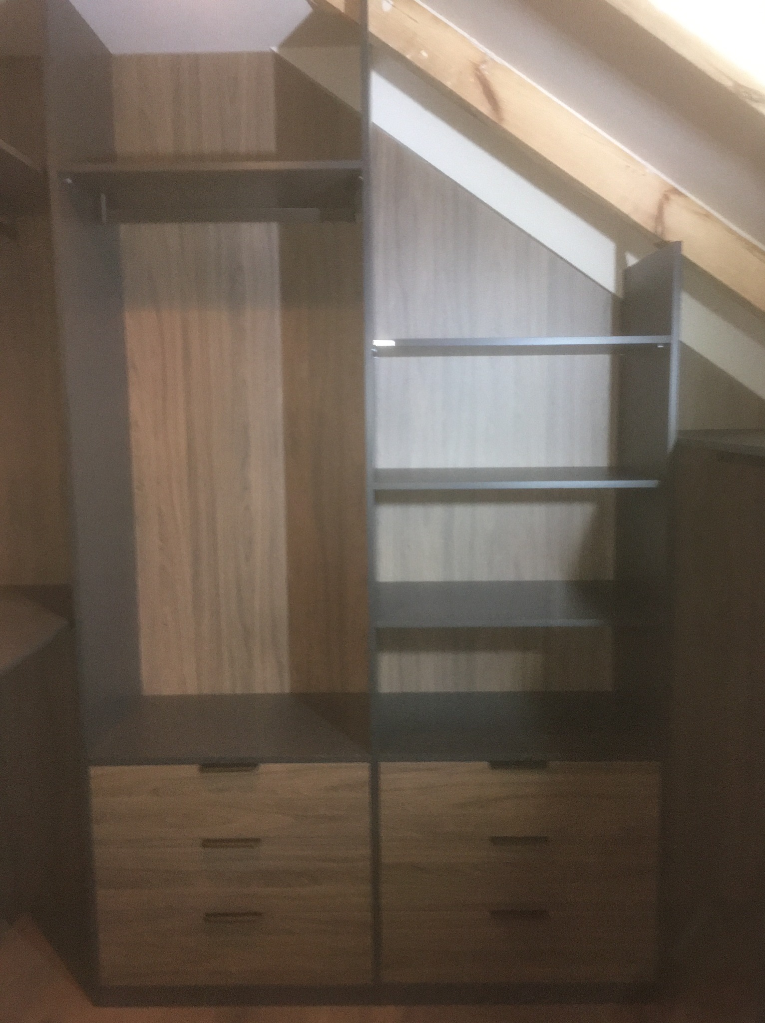 Drewniana szafa wnękowa z szufladami i półkami, dopasowana do skosu dachu. Widoczne elementy montażowe i wykończenie.