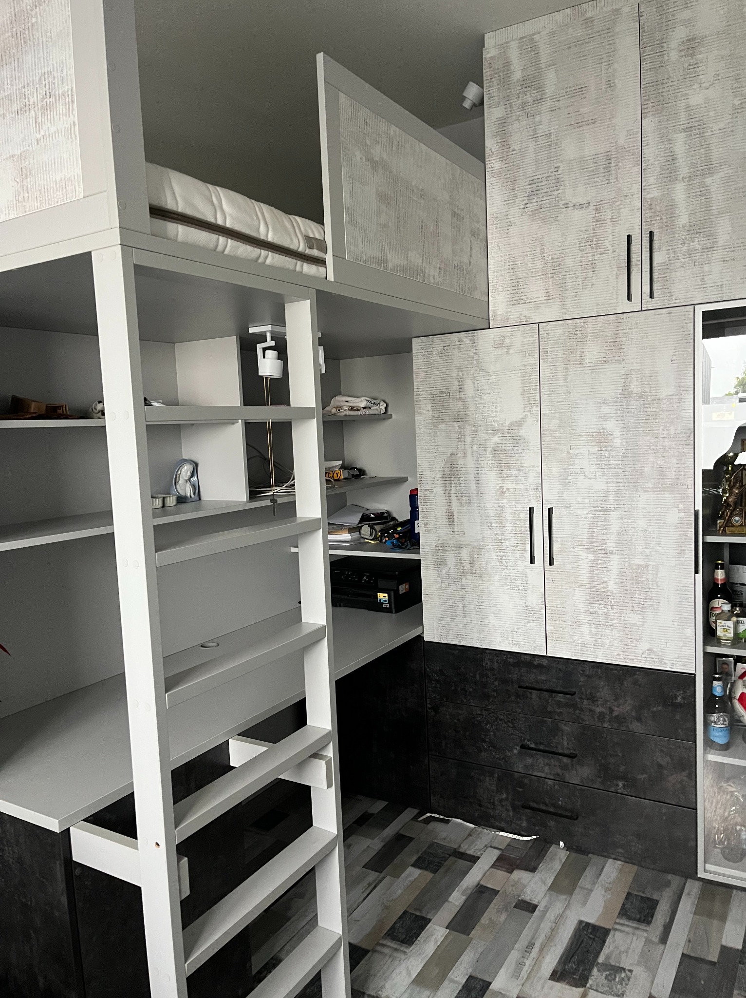 Funkcjonalna zabudowa pokoju młodzieżowego z łóżkiem na antresoli, biurkiem i szafami. Biało-szara kolorystyka, nowoczesny design. Widoczna drabinka i półki.