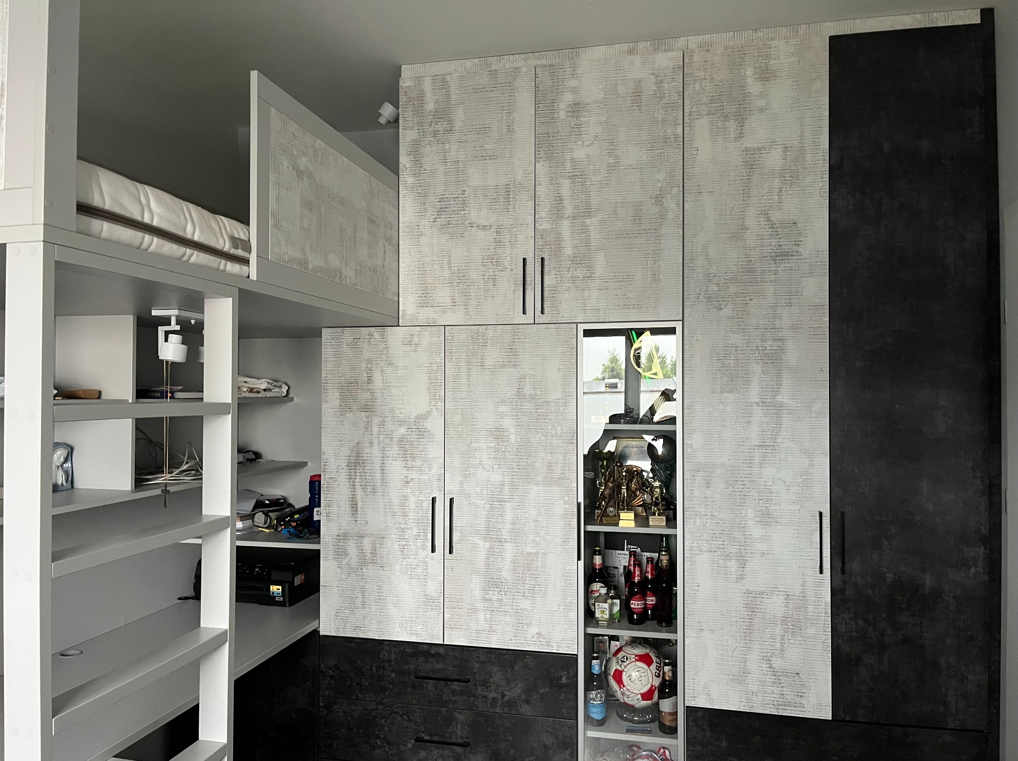 Funkcjonalna szafa na wymiar z łóżkiem na antresoli, jasne fronty z delikatnym wzorem, czarne akcenty, półki z widocznymi przedmiotami i trofeami sportowymi.