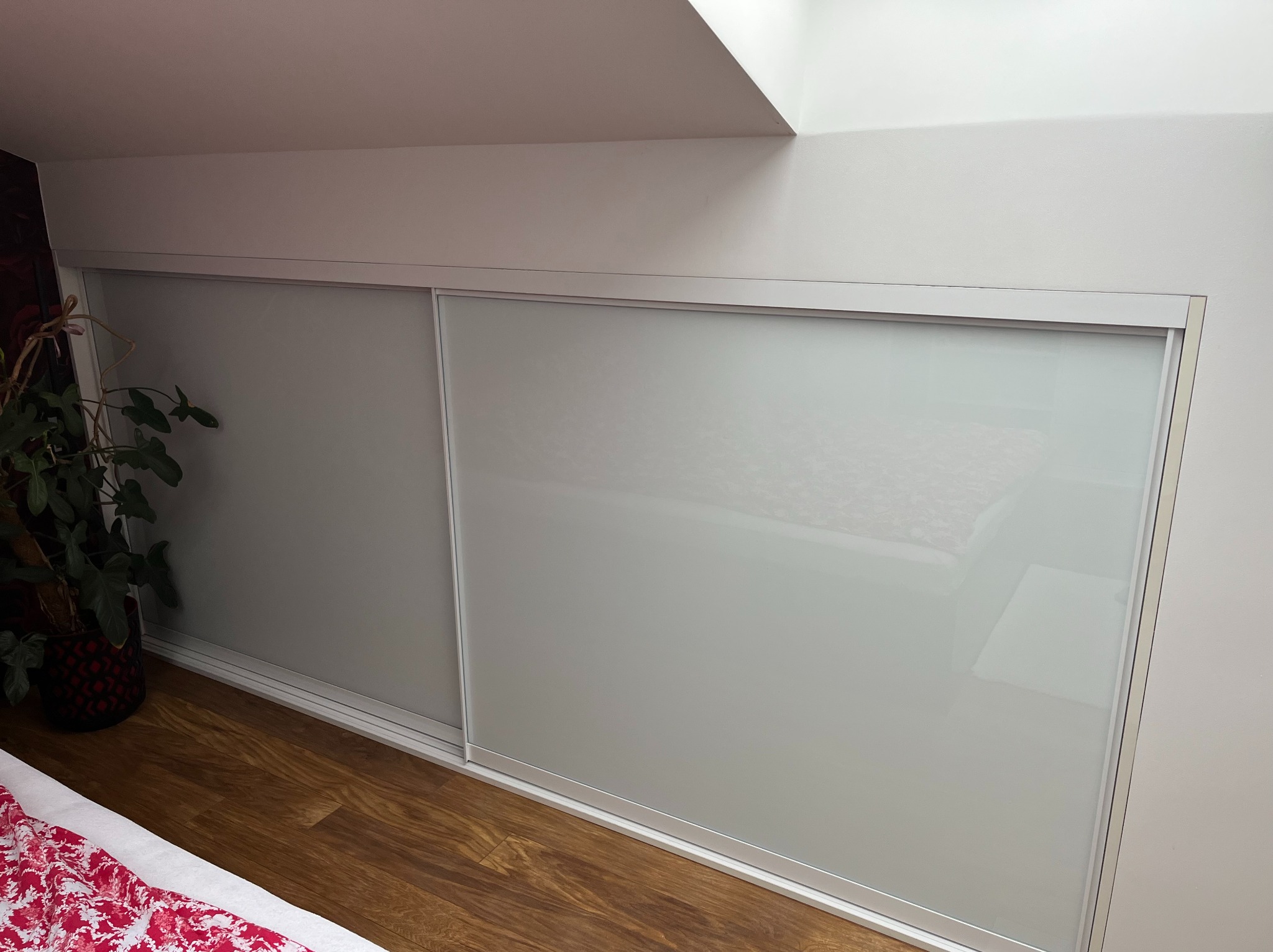 Biała szafa wnękowa z przesuwnymi drzwiami z mlecznego szkła, dopasowana do skosu dachu, z rośliną doniczkową obok i drewnianą podłogą.