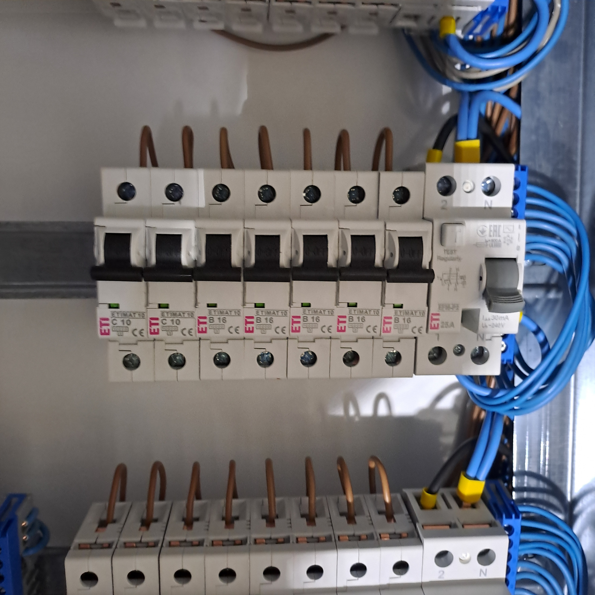 Szczegółowe ujęcie tablicy rozdzielczej z zainstalowanymi bezpiecznikami ETI i niebieskimi przewodami. Widoczne oznaczenia C10 i B16 na poszczególnych elementach.