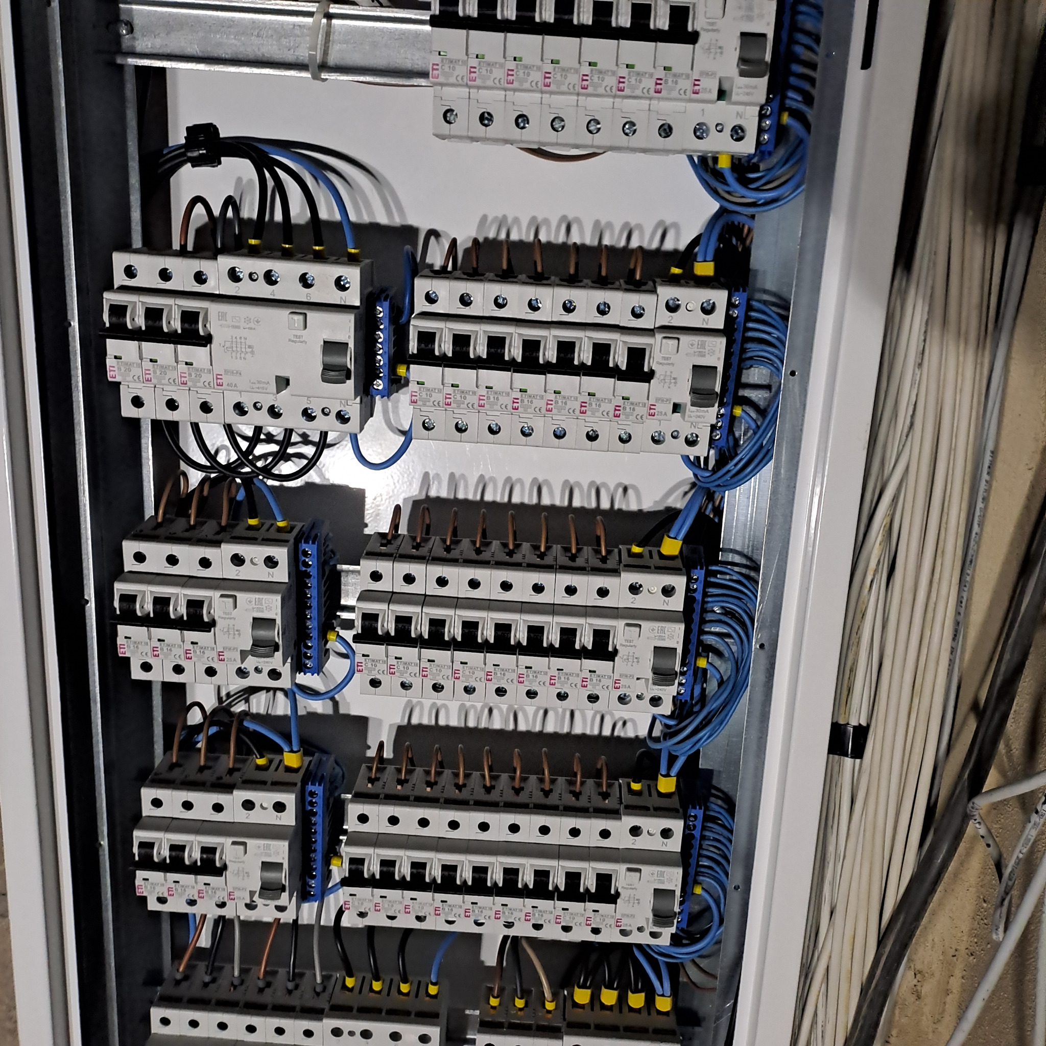 Rozdzielnica elektryczna z bezpiecznikami i okablowaniem w kolorach niebieskim, czarnym i brązowym, zamontowana w białej obudowie. Widoczne przewody elektryczne.