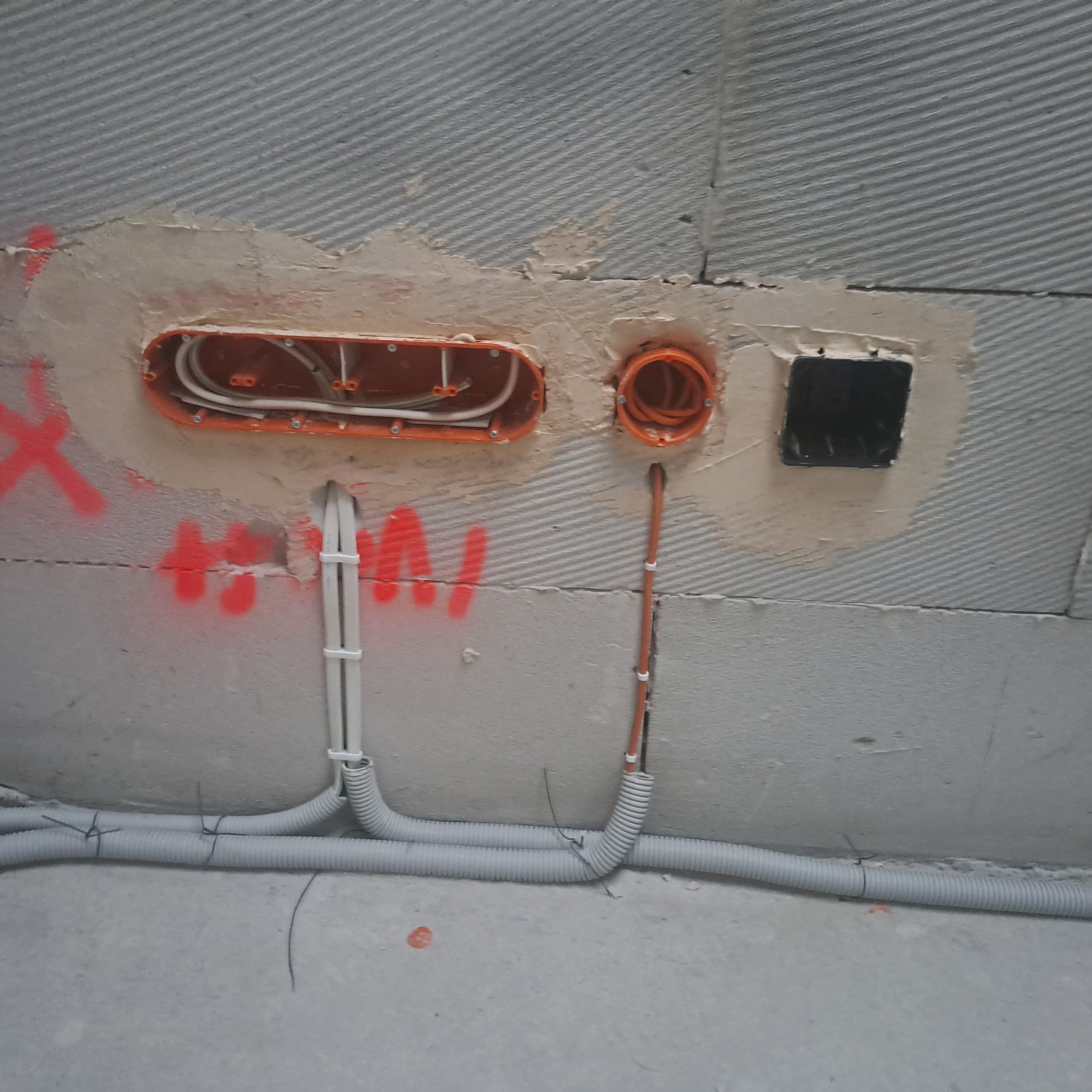 Surowy stan instalacji elektrycznej: puszki podtynkowe (pomarańczowe i czarna) osadzone w ścianie z szarego bloczka, widoczne przewody i peszle. Ślady czerwonej farby.