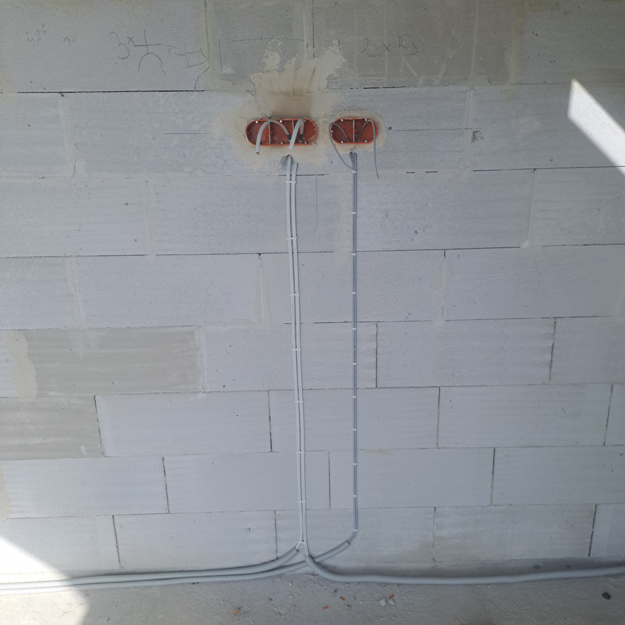 Dwa przewody elektryczne w białych peszlach, wychodzące z pomarańczowych puszek w ścianie z betonu komórkowego. Widoczne kable i oznaczenia na ścianie.