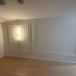 Strefa Remontu - Białe ściany z dekoracyjnymi listwami i oświetleniem punktowym w pustym pokoju z drewnianą podłogą. Minimalistyczny design wnętrza.