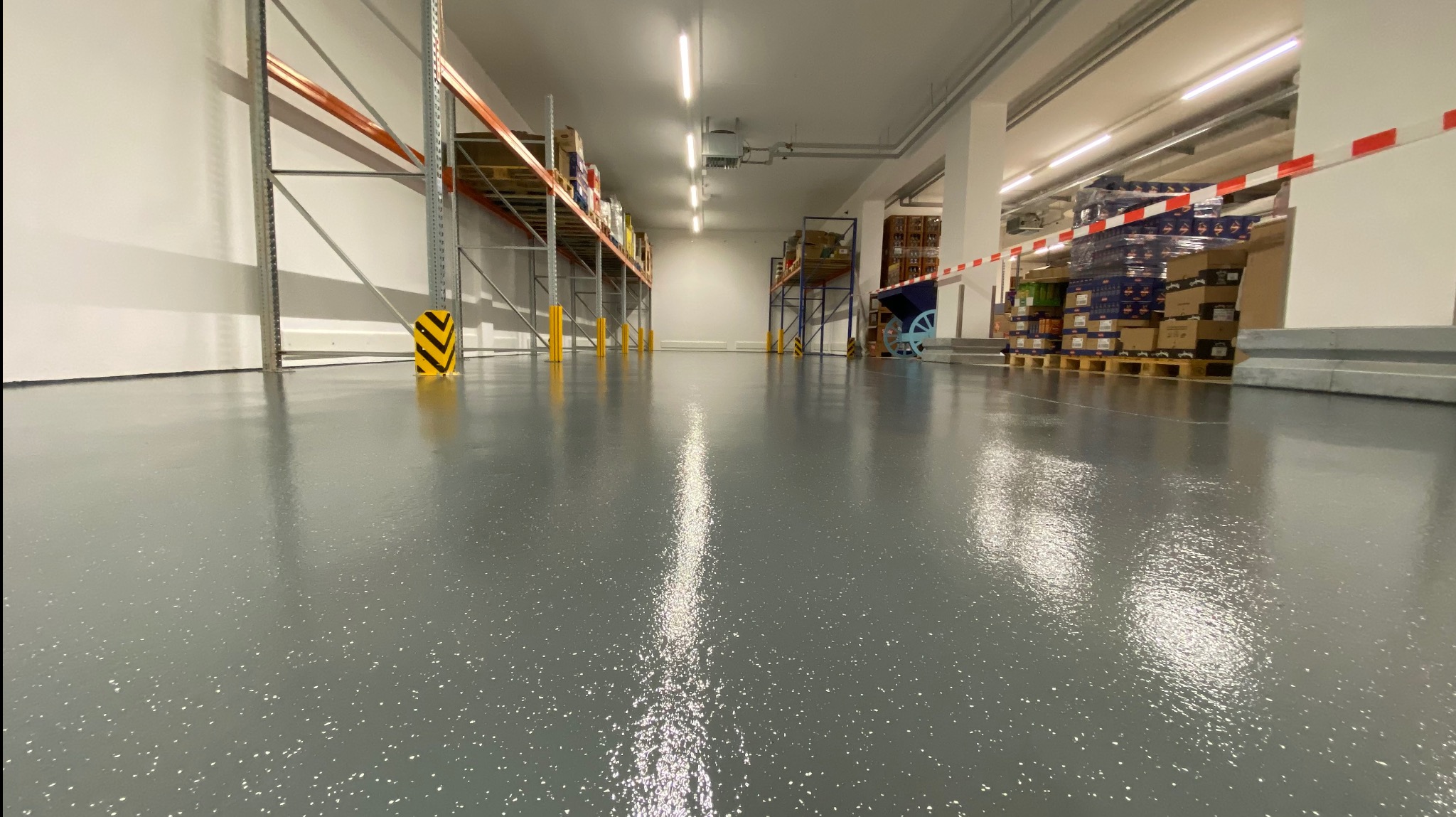 Błyszcząca, szara posadzka epoksydowa w magazynie z metalowymi regałami. Podłoga z delikatnym wzorem, dobrze oświetlona, z żółto-czarnymi osłonami słupów.