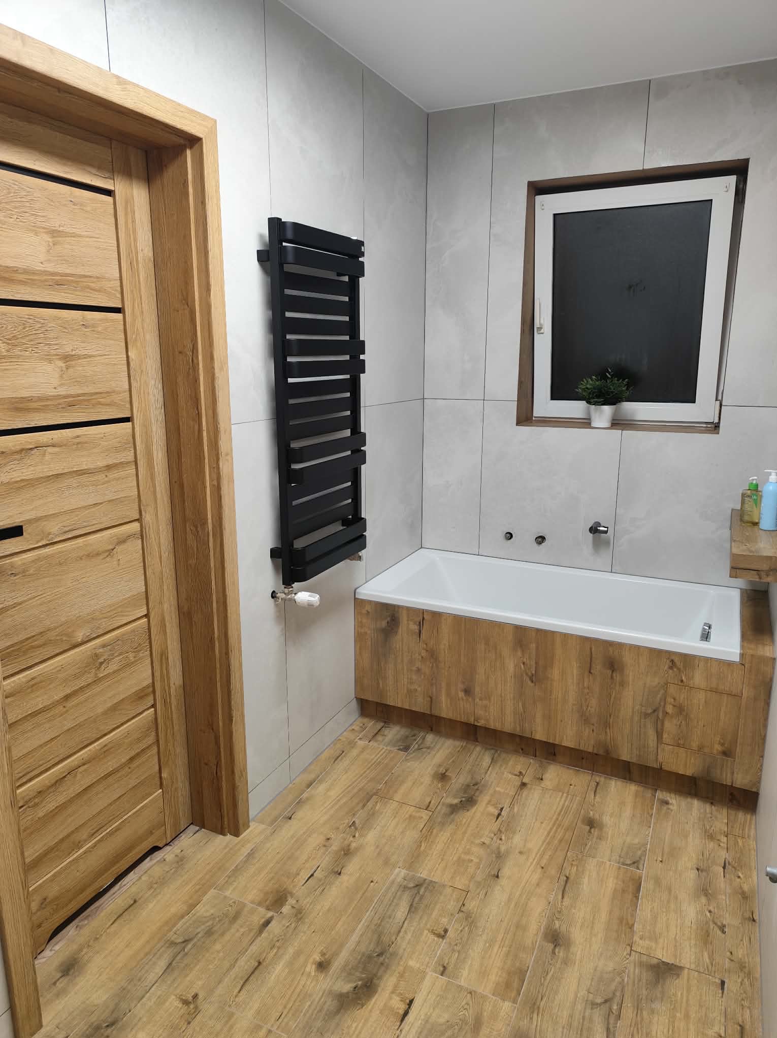 Nowoczesna łazienka po remoncie: drewnopodobne płytki, zabudowana wanna, grzejnik drabinkowy i drewniane drzwi. Jasne płytki na ścianach i okno z rośliną.