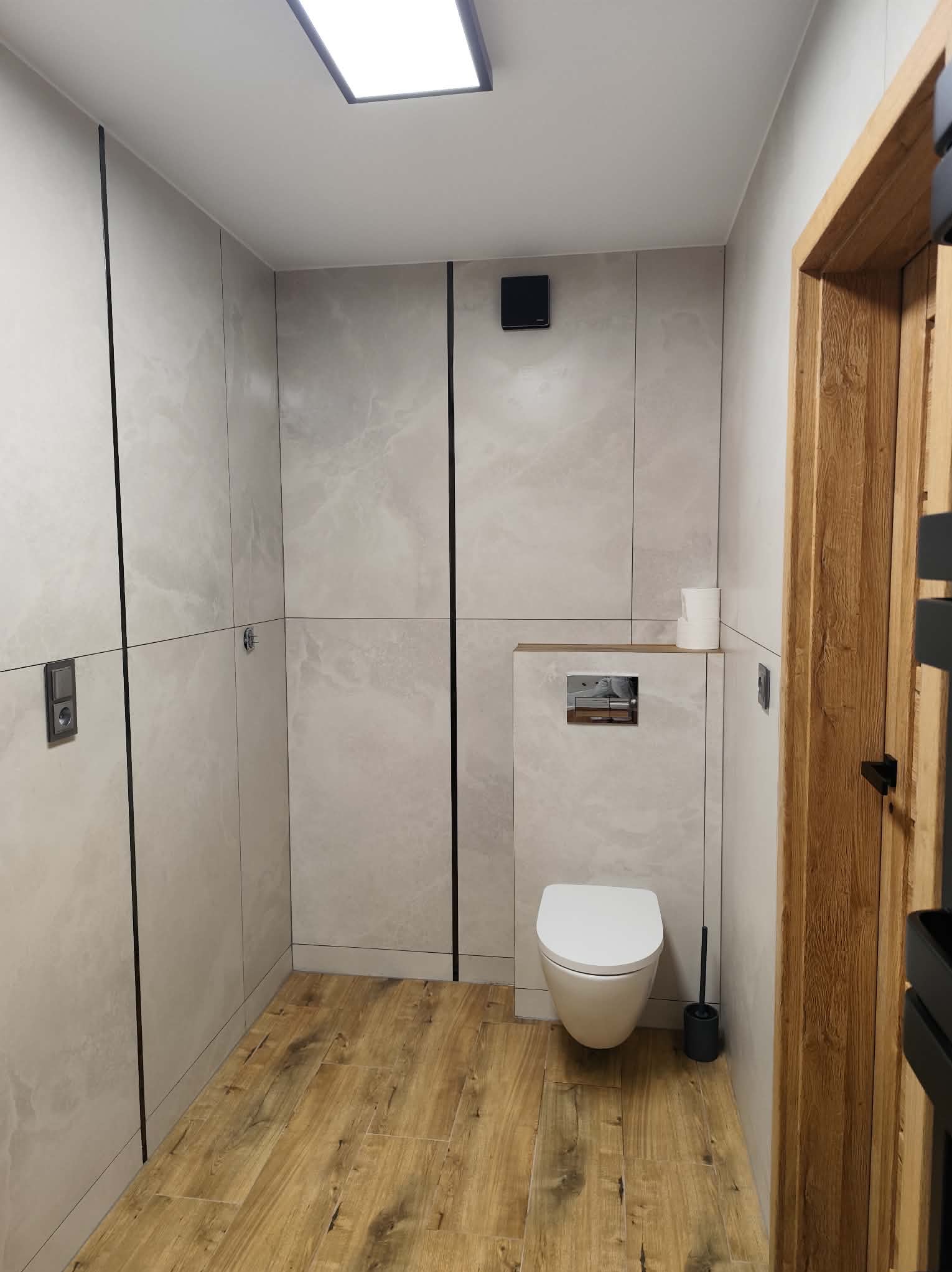 Nowoczesna łazienka z drewnianą podłogą i płytkami imitującymi marmur, akcentowana czarnymi listwami. Wisząca toaleta i drewniana rama drzwi dodają elegancji.
