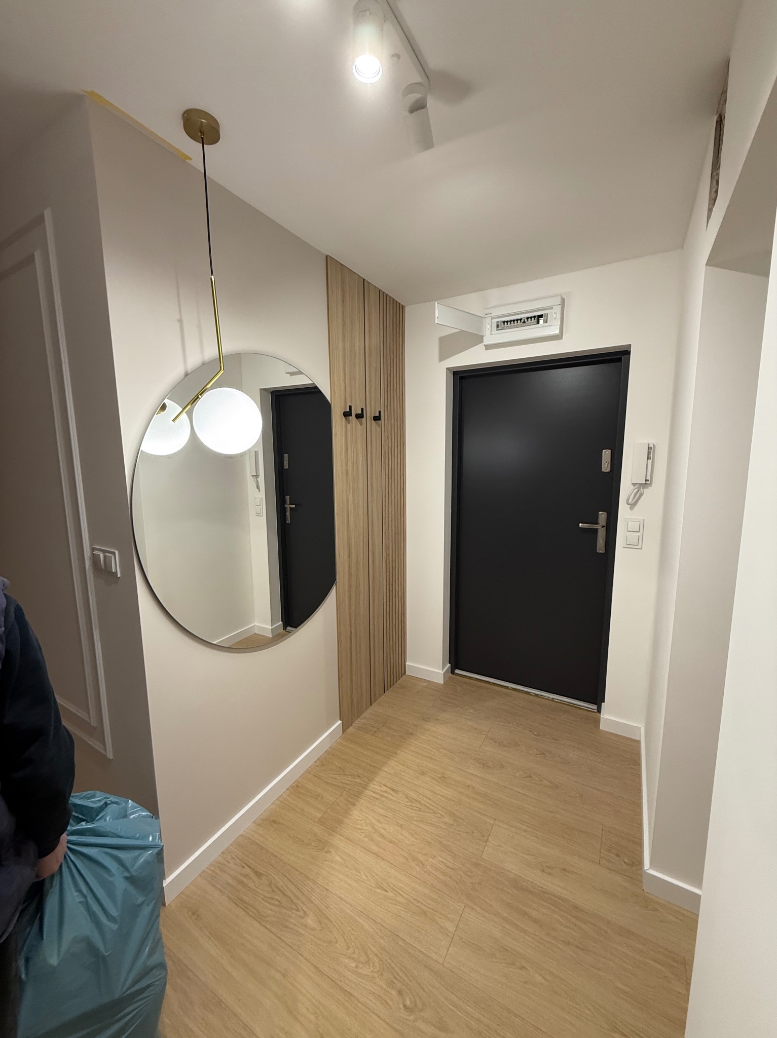 Nowoczesny przedpokój po remoncie: jasne ściany, drewniana podłoga, okrągłe lustro z lampą i czarne drzwi wejściowe. Minimalistyczny design z elementami dekoracyjnymi.