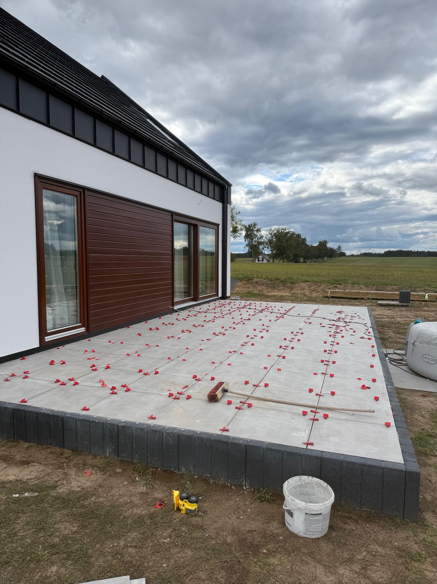Układanie jasnoszarych płytek na tarasie domu jednorodzinnego z użyciem czerwonych krzyżyków dystansowych. Widoczne narzędzia i wiadro. Tło: pochmurne niebo i pole.