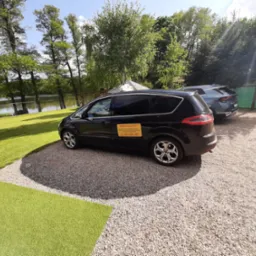 Czarny van taxi z żółtą naklejką informacyjną parkuje na żwirowym podłożu z widokiem na jezioro i zieleń w tle. Na szybie numer telefonu do zamówienia.