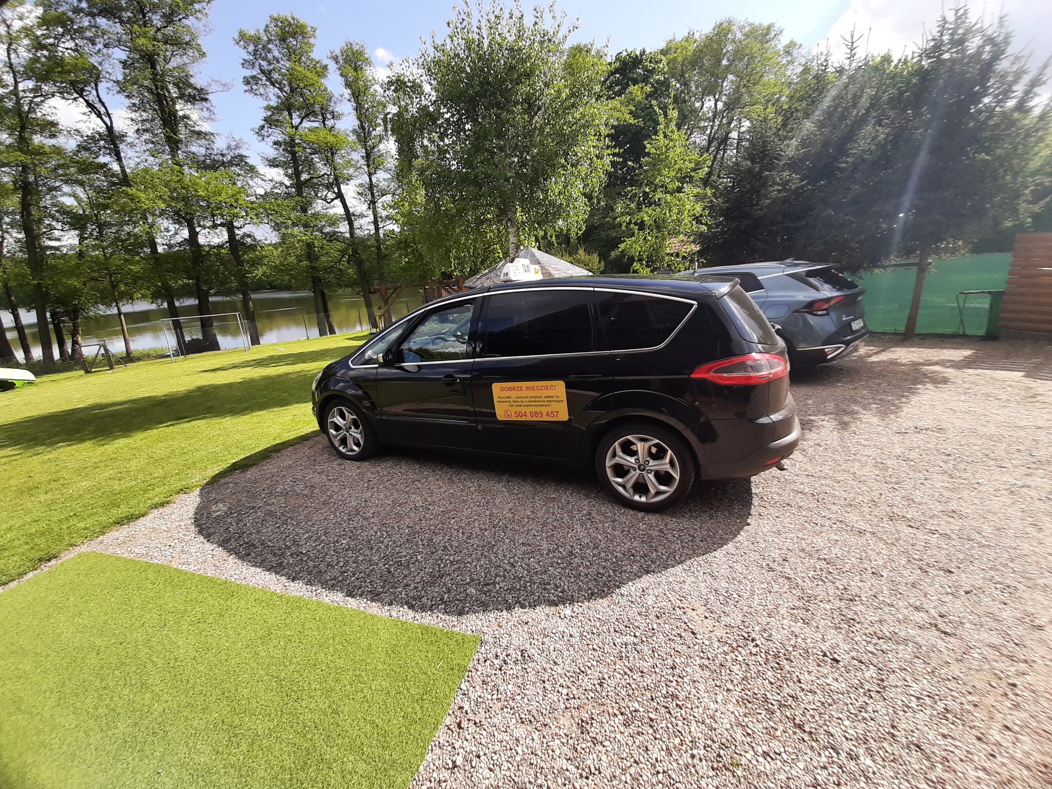 Czarny van taxi z żółtą naklejką informacyjną parkuje na żwirowym podłożu z widokiem na jezioro i zieleń w tle. Na szybie numer telefonu do zamówienia.