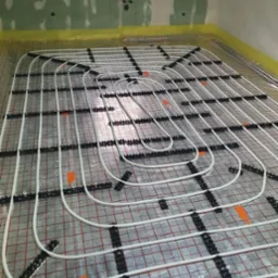 Białe rury ogrzewania podłogowego, mocowane czarnymi uchwytami do folii z czerwoną siatką na tle surowej ściany. Widoczny etap instalacji.