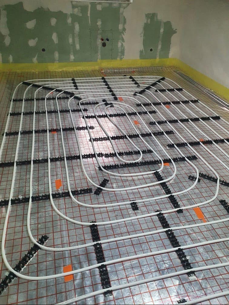 Białe rury ogrzewania podłogowego, mocowane czarnymi uchwytami do folii z czerwoną siatką na tle surowej ściany. Widoczny etap instalacji.