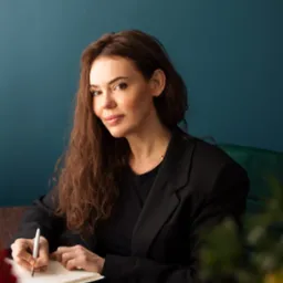 Kobieta w czarnej marynarce pisze w notatniku, Warszawa. Uśmiechnięta brunetka z długimi włosami, elegancki strój, notatnik i długopis na biurku. Neutralne tło.