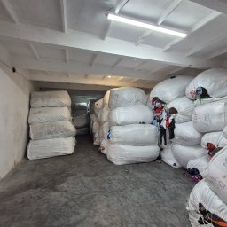 Rafal Żołna - Magazyn z workami odzieży używanej, ułożonymi w stosy pod betonowym sufitem z lampą. Widoczne napisy na workach i fragmenty ubrań.