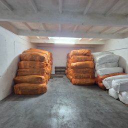 Rafal Żołna - Magazyn z workami odzieży używanej w pomarańczowych i białych workach, ułożonymi w stosy na betonowej podłodze w pomieszczeniu z betonowym sufitem.