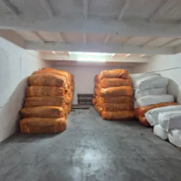 Magazyn z workami odzieży używanej w pomarańczowych i białych workach, ułożonymi w stosy na betonowej podłodze w pomieszczeniu z betonowym sufitem.