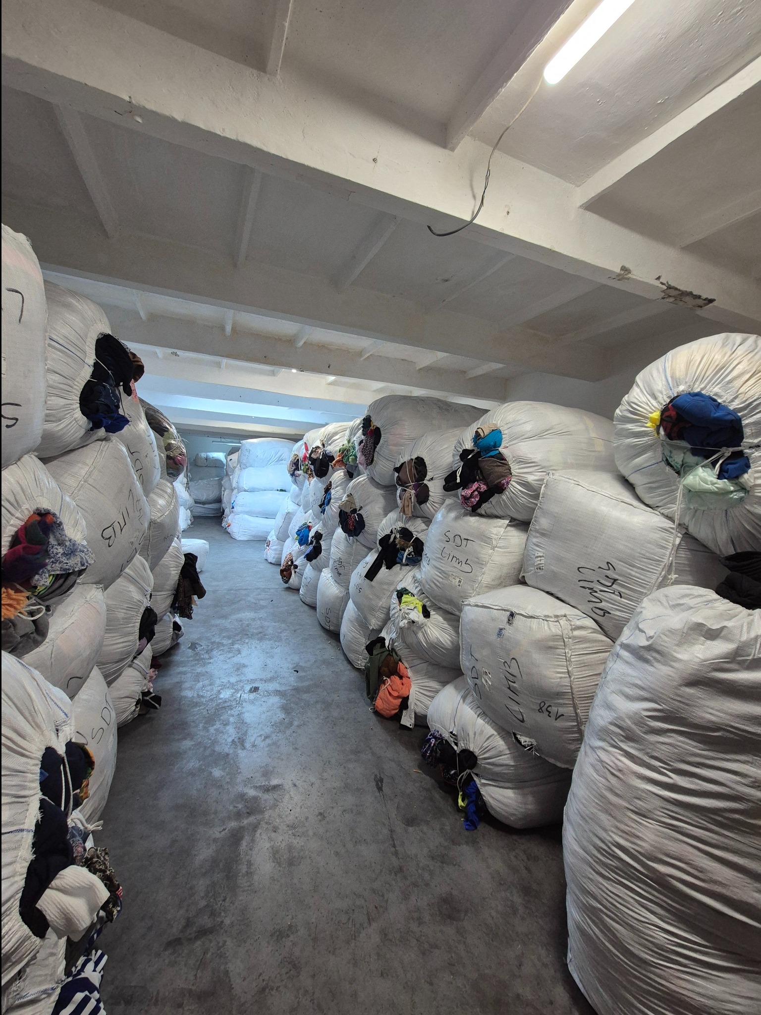 Magazyn z workami odzieży używanej; worki ułożone w rzędach, widoczne fragmenty ubrań. Szare betonowe podłoże, biały sufit z lampami.