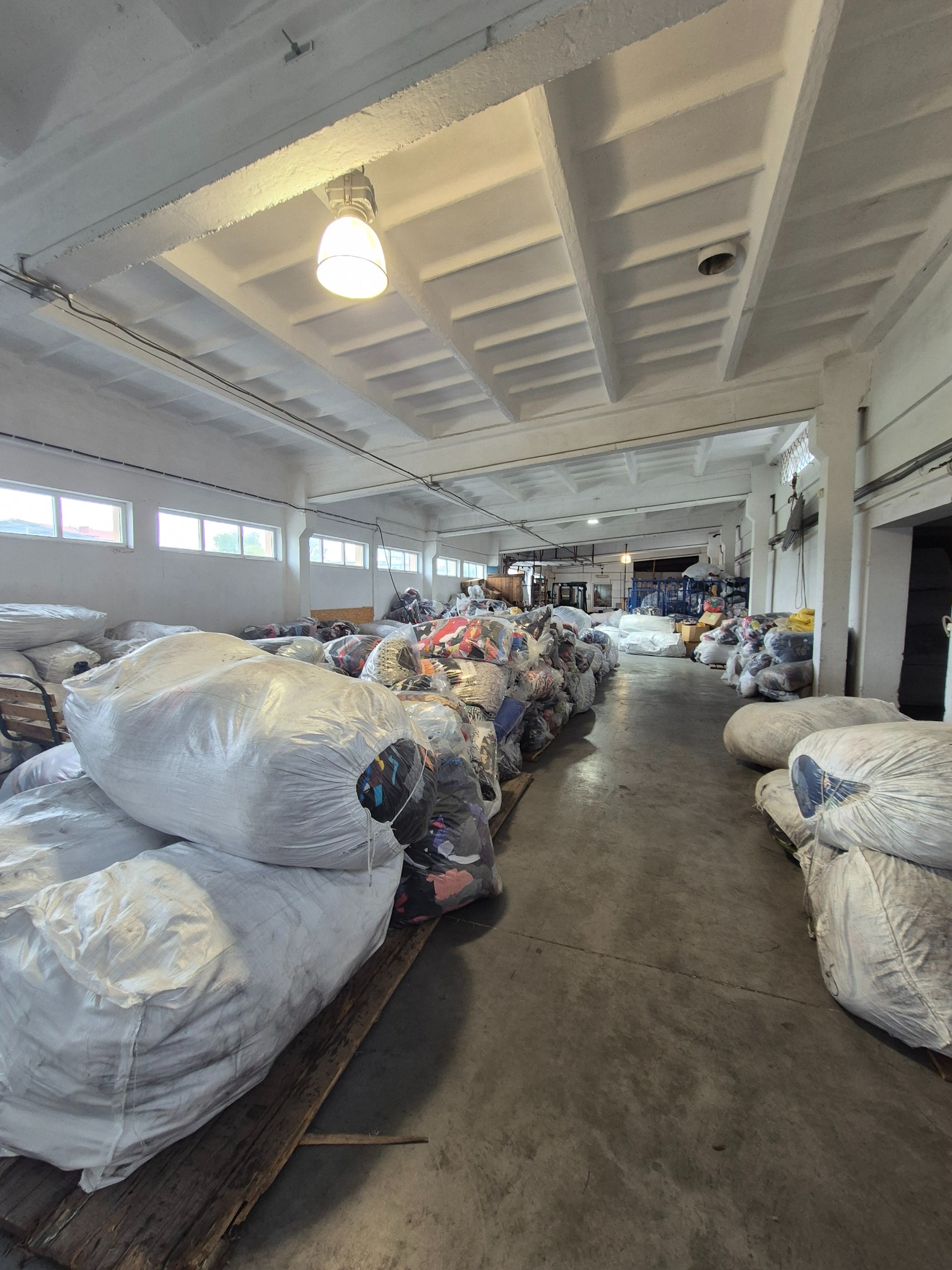 Magazyn z workami odzieży używanej, widok w głąb hali. Worki ułożone w rzędach na paletach. Białe ściany, sufit z betonowymi belkami, okna w górnej części ścian.