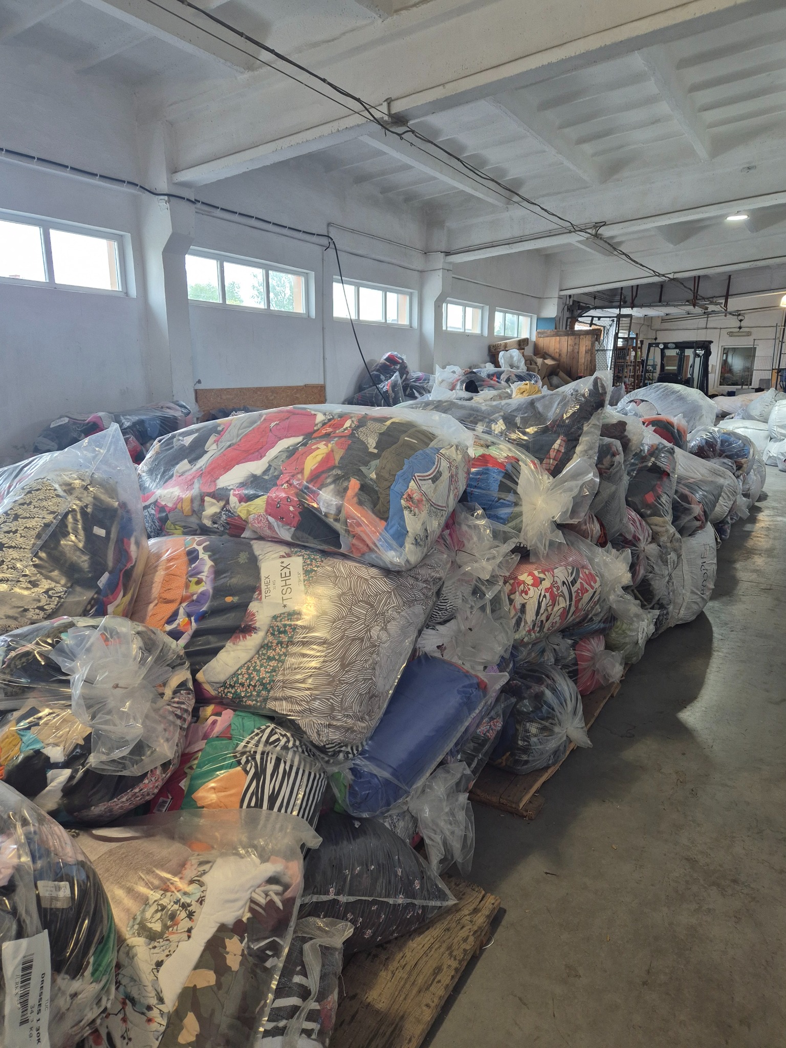 Magazyn z paletami worków wypełnionych tekstyliami, widoczne etykiety i faktura różnych materiałów. Surowe, przemysłowe wnętrze.