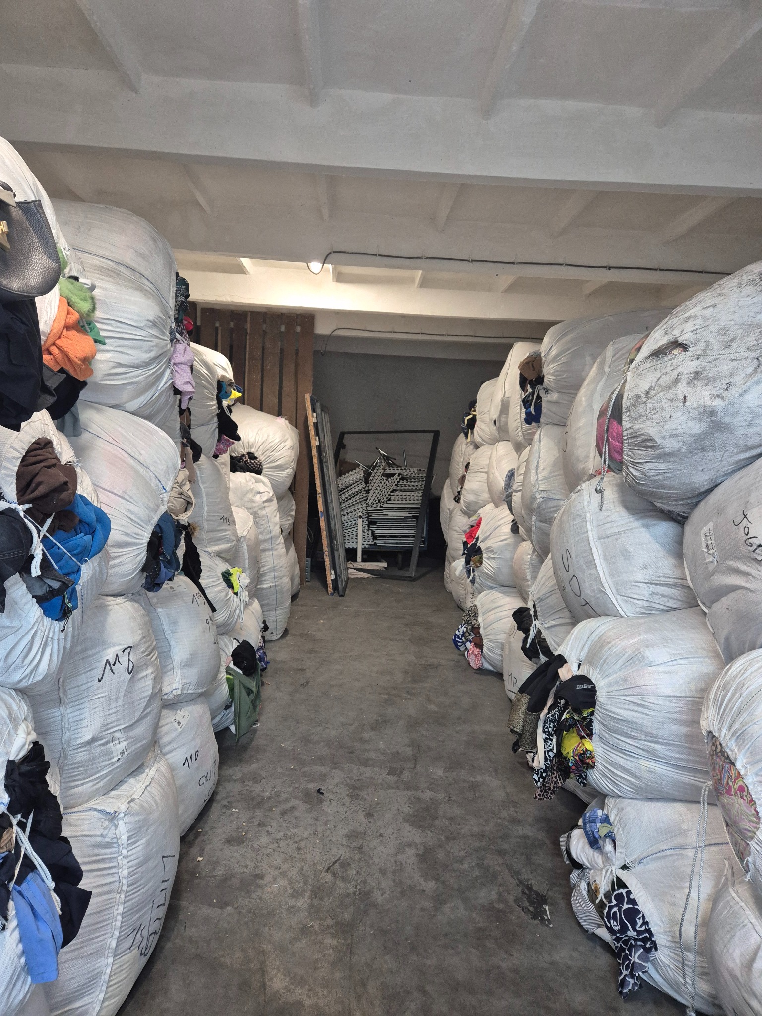Magazyn z workami odzieży używanej; widoczne worki z napisami, częściowo wystająca odzież, betonowa podłoga i sufit.