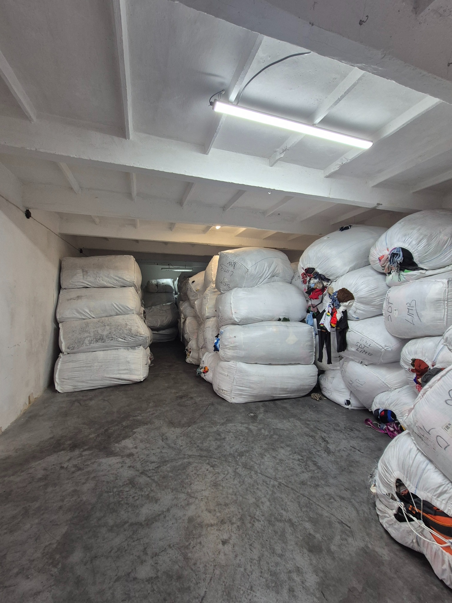 Magazyn z workami odzieży używanej, ułożonymi w stosy pod betonowym sufitem z lampą. Widoczne napisy na workach i fragmenty ubrań.