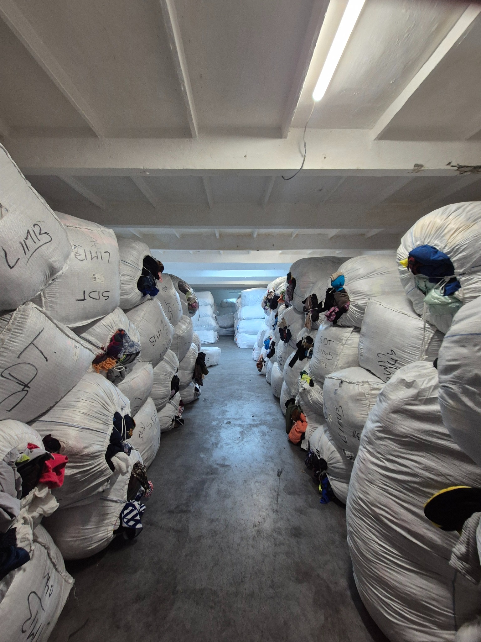 Rzędy worków z odzieżą w magazynie, widoczne fragmenty ubrań, betonowa podłoga i sufit z lampami. Ujęcie z perspektywy alejki między workami.