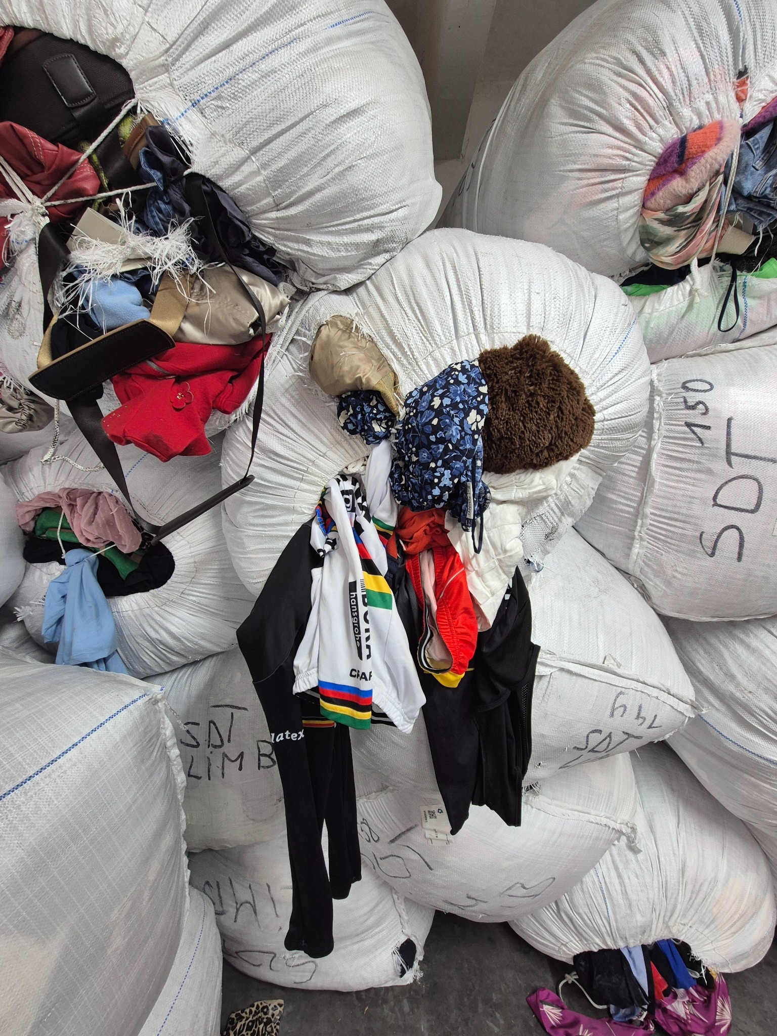 Stos worków z odzieżą. Białe, spiętrzone worki z widoczną zawartością, w tym ubrania w różnych kolorach i fasonach. Na workach widoczne napisy.
