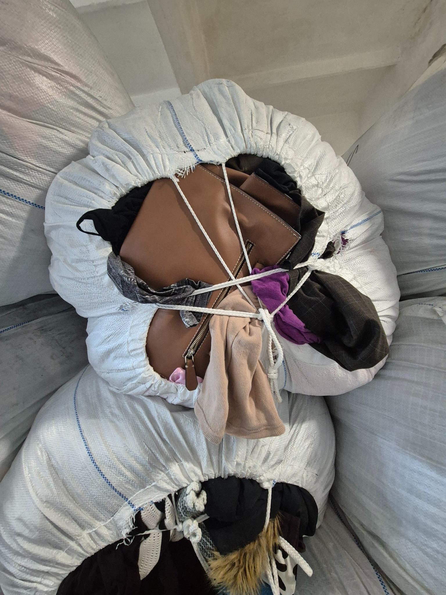 Wypełniony worek z odzieżą, przewiązany sznurkiem, z widoczną brązową torebką, fragmentami ubrań i suwakiem. W tle inne worki z odzieżą.